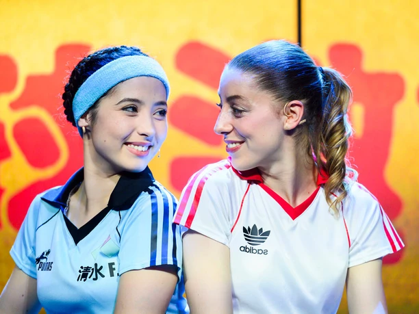Kick Like a Woman - Ein Musical über den Mut, für Träume zu kämpfen Zwei Fußballerinnen in Trikots lächeln sich vor gelb-roter Bühnenkulisse an