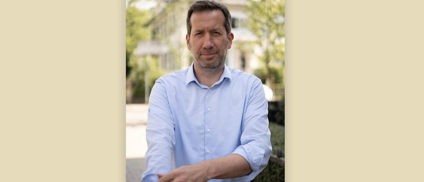 Marc Raschke im hellblauen Hemd vor unscharfem Gebäude und Grün.