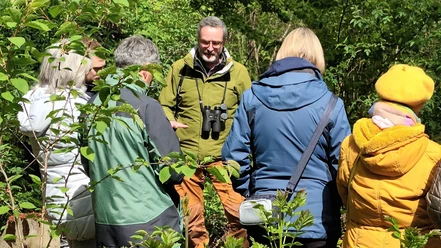 Gruppe bei einer NABU-Naturführung im Grünen, Leiter mit Fernglas erklärt die Umgebung.