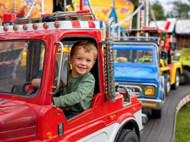 Lächelnder Junge sitzt in einem roten Spielzeugauto auf einem Jahrmarkt. Im Hintergrund weitere bunte Autos.