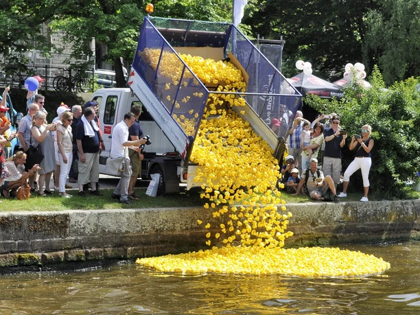 A truck dumping a load of yellow rubber ducks into a river, while a group of people
