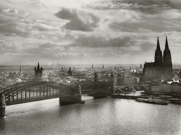 A historic black-and-white view of Cologne featuring the Hohenzollern Bridge, the cathedral, and the Rhine