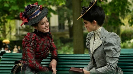 Zwei Frauen in historischen Kleidern sitzen lächelnd auf einer Parkbank im Grünen.