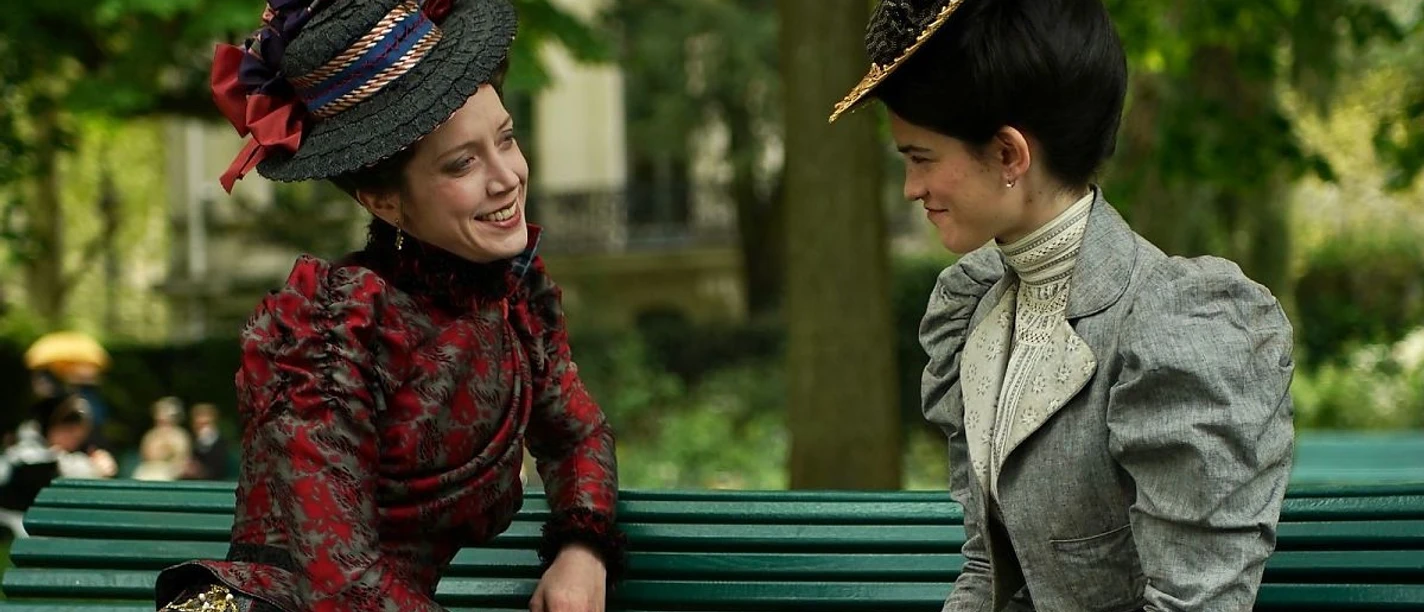 Zwei Frauen in historischen Kleidern sitzen lächelnd auf einer Parkbank im Grünen.