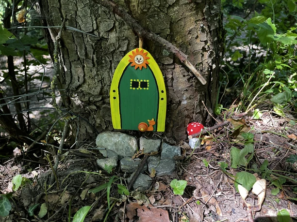 Eine kleine grün-gelbe Wichteltür mit Schnecke und Sonne, ist an der Rinde eines Baumes befestigt. Unterhalb der Tür befinden sich kleine Steine als Eingang.