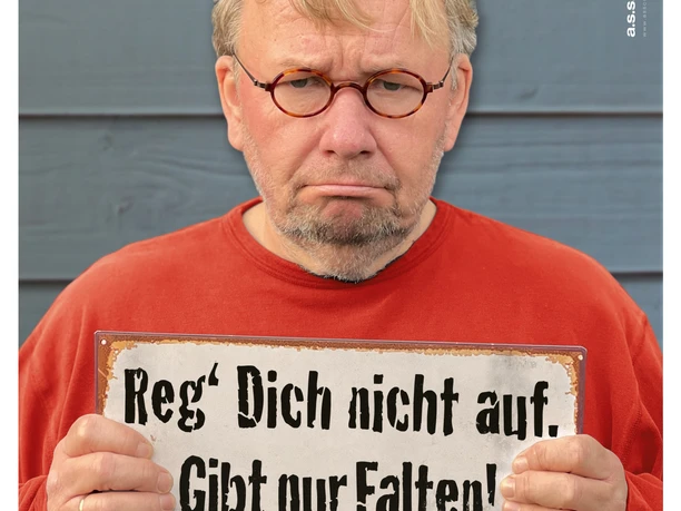Bernd Stelter mit ernster Miene hält ein Schild: Reg’ Dich nicht auf. Gibt nur Falten!