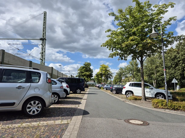 Parkplatz am Bahnhof Rastede mit Autos, Bäumen und Bahn-Oberleitung unter Wolken.