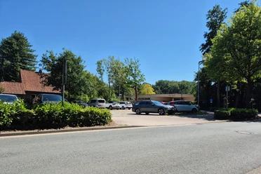 Parkplatz am Freibad Rastede mit Autos, Hecken und Bäumen bei klarem Himmel.