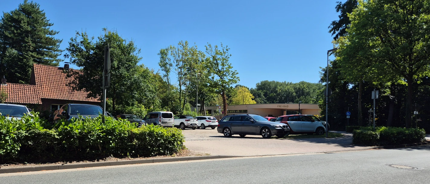 Parkplatz am Freibad Rastede mit Autos, Hecken und Bäumen bei klarem Himmel.