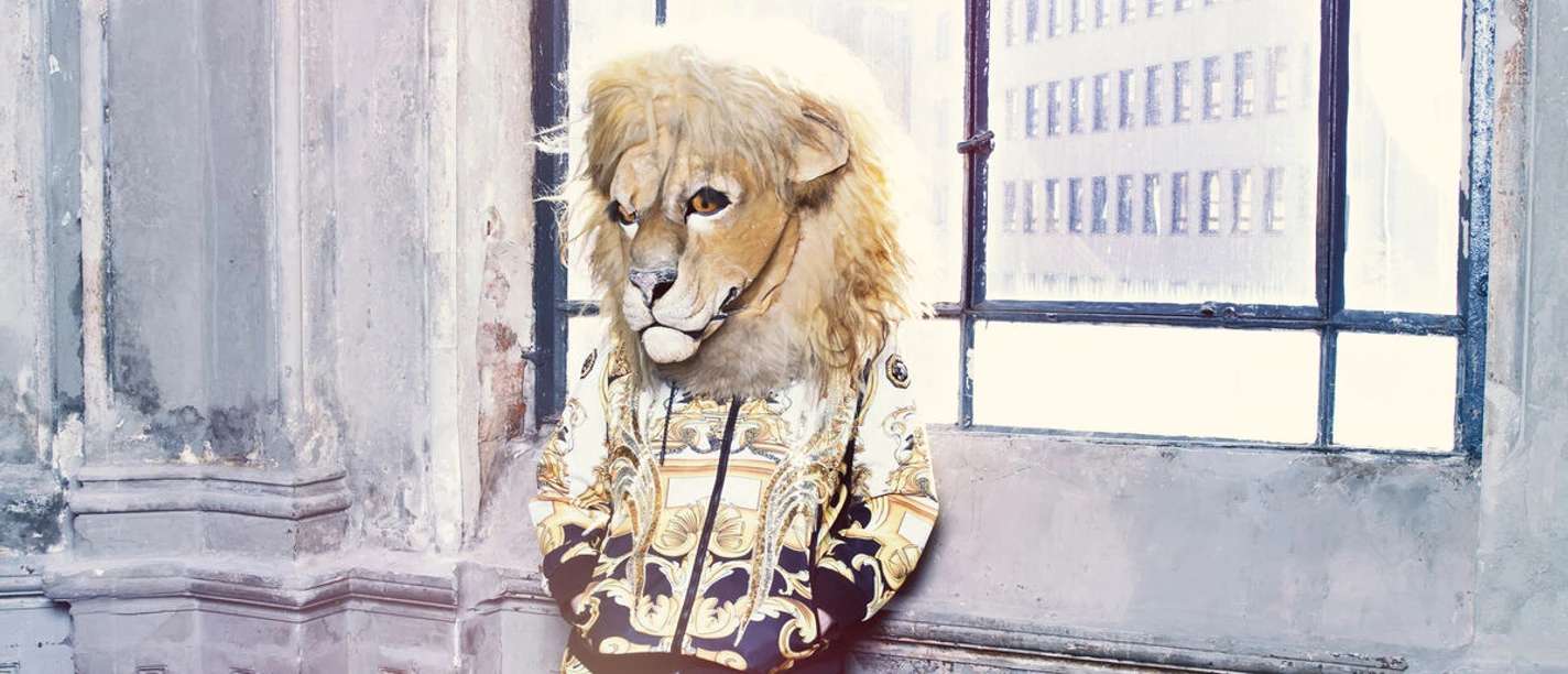 Bühnenfigur mit Löwenmaske in gold-schwarzem Outfit vor hohem Fenster im Altbau