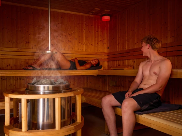 Zwei junge Personen nutzen die Sauna, die Dame liegt auf der saunabank, der Herr sitzt auf der Saunabank und unterhält sich mit der Dame