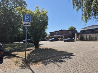 Öffentlicher Parkplatz in Rastede mit blauem Parkschild, Bäumen, Autos und Wohngebäuden.