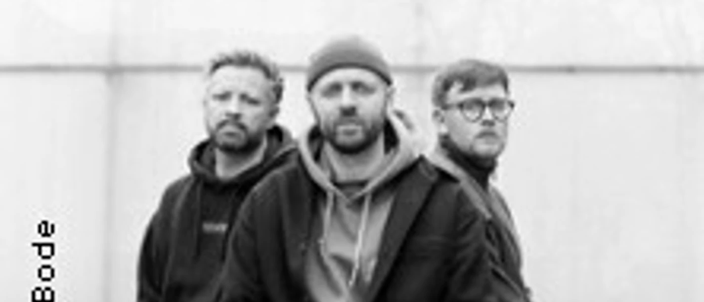 Schwarz-Weiß-Porträt von drei sitzenden Musikern vor heller Betonwand