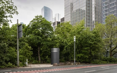 Außenansicht einer dunkelgrauen Säule, im Hintergrund Bäume und moderne Hochhäuser.