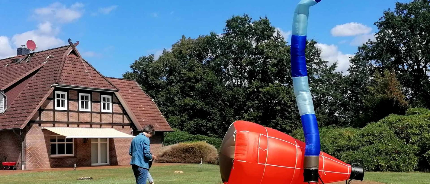 Rote Luftskulptur mit blauem Schlauch im Garten vor einem Fachwerkhaus.