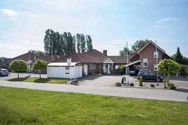 Backsteinhotel am Deich mit Parkplatz, Rasenfläche und Bäumen bei sonnigem Wetter.