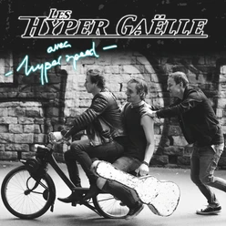 Drei Männer mit Moped und Gitarrenkoffer vor einer Steinmauer.