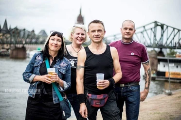 Four people with drinks on the riverbank in front of a bridge.