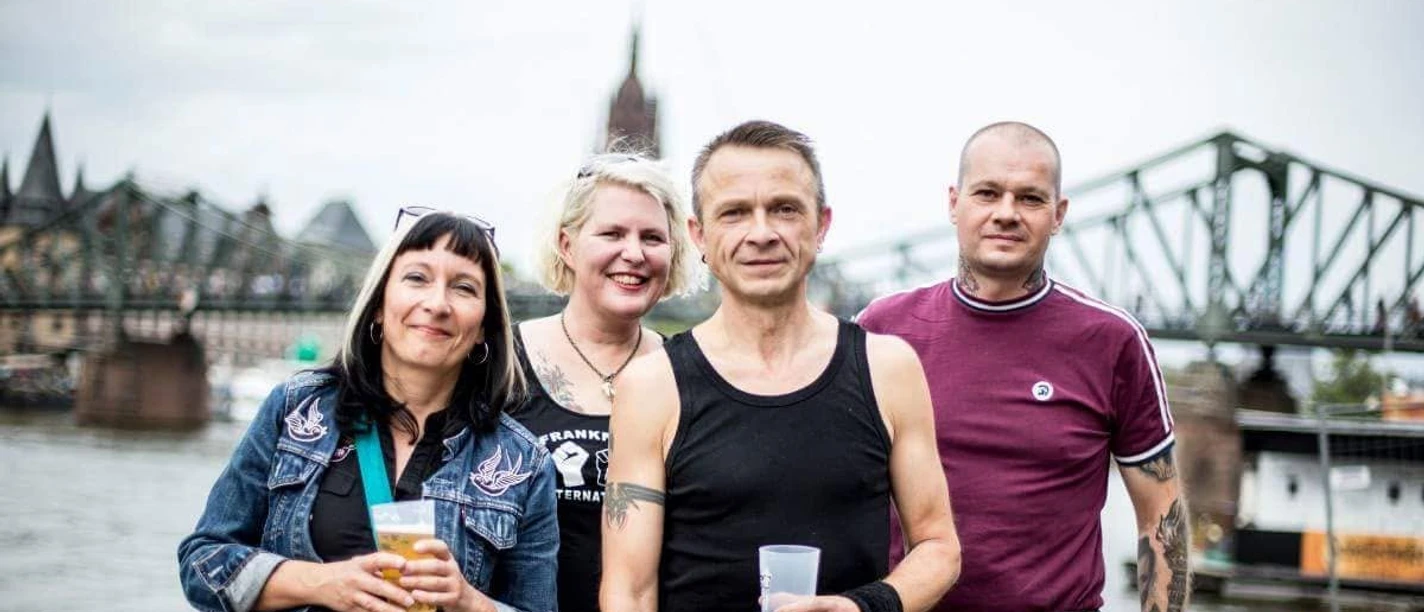 Four people with drinks on the riverbank in front of a bridge.