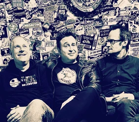 Three men sitting in front of a wall covered with stickers.