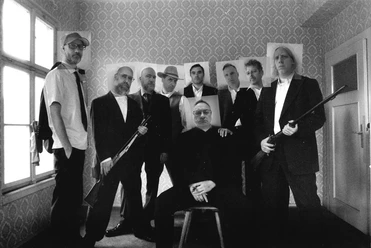 Nine men in suits, some of them armed, in a room.