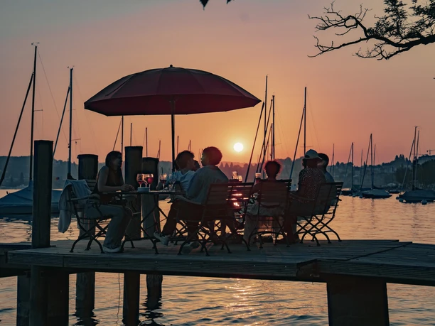 Guests at Sunset Bar sit on a lakeside jetty at dusk, with boats silhouetted behind them