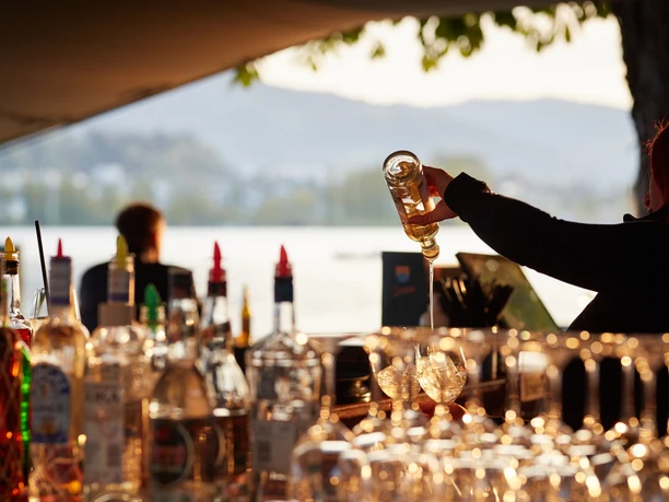 Bartender pours a drink at Sunset Bar, with bottles glowing before a lakeside view.