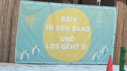 Banner am Beachfeld mit Schriftzug „Rein in den Sand und los geht’s!“ und Strandbällen.
