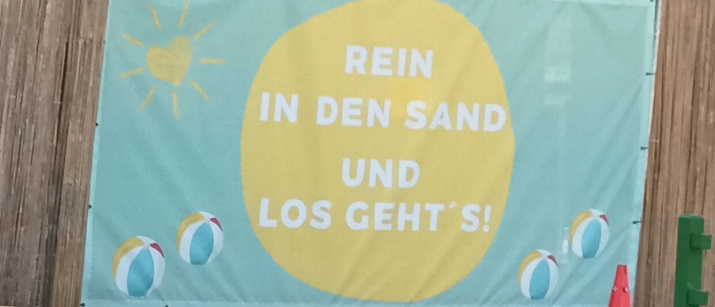 Banner am Beachfeld mit Schriftzug „Rein in den Sand und los geht’s!“ und Strandbällen.