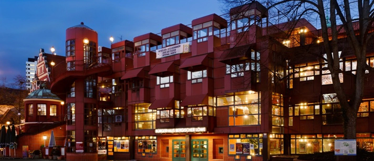 Abendansicht des Bürgerhauses Bergischer Löwe mit beleuchteter roter Fassade