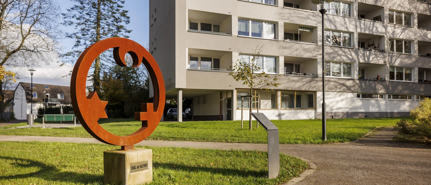 Rostrote Skulptur „Engel der Kulturen“ auf einer Wiese vor einem Wohnhaus