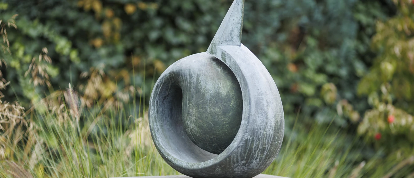 Abstrakte Metallskulptur „aus sich heraus“ auf Steinsockel vor grünem Bewuchs