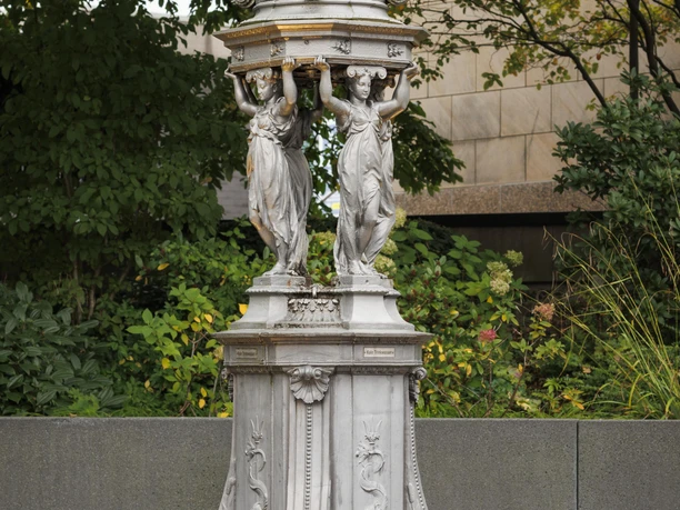 Silberner Wallace-Brunnen mit Frauenfiguren und Kuppeldach vor grünem Stadtgarten