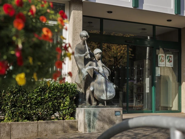 Bronzeskulptur „Musizierende Kinder“ vor einem Eingang, daneben rote Blumen