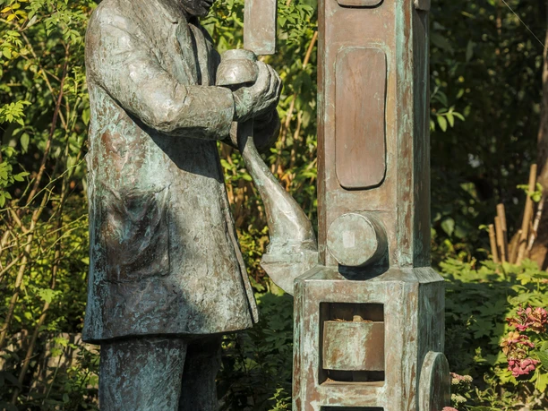 Bronzeskulptur eines Schuhmachers an einer Maschine, umgeben von Grün