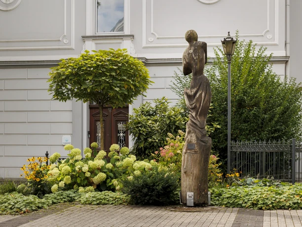 Holzskulptur „Peterhead“ in einem bepflanzten Vorgarten vor heller Fassade
