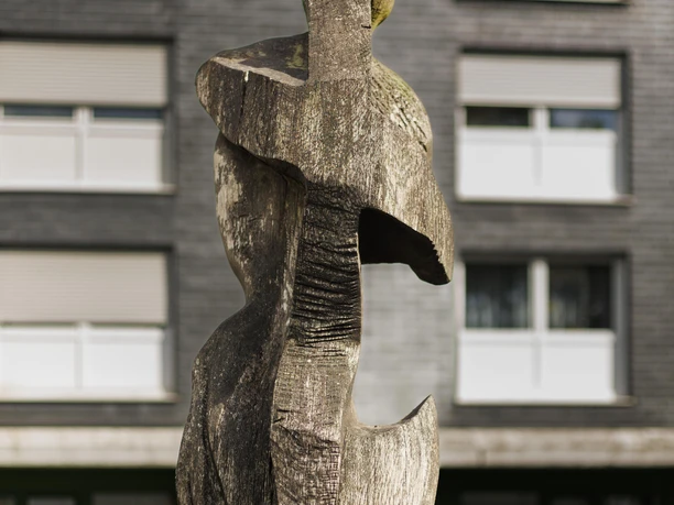 Abstrakte Holzskulptur „Peterhead“ vor grauem Wohnhaus in Wermelskirchen