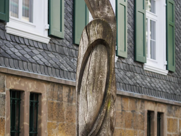 Holzskulptur Metamorphose vor Schieferfassade mit grünen Fensterläden