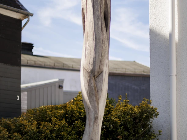 Hohe Holzskulptur vor Hecke und Hausfassade unter hellem Himmel in Wermelskirchen