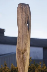 Hohe Holzskulptur mit Rissen und Öffnung vor unscharfem Gebäude und Sträuchern