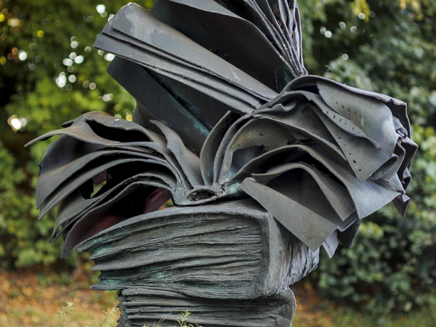 Skulptur „Bücherstapel“ mit aufgeschlagenen Metallbüchern vor grünem Parkhintergrund