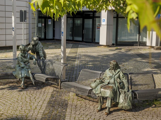 Bronzeskulptur „Begegnung“ mit drei Figuren auf Bänken vor einem Gebäude in Wermelskirchen