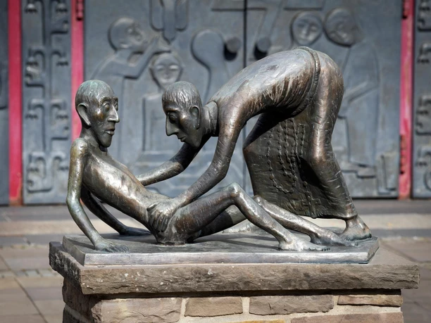 Bronzeskulptur mit gebeugter und sitzender Figur vor Kirchenportal in Marienheide