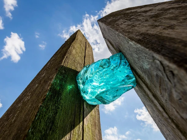 Türkis leuchtender Glaskörper zwischen Holzstelen vor blauem Himmel