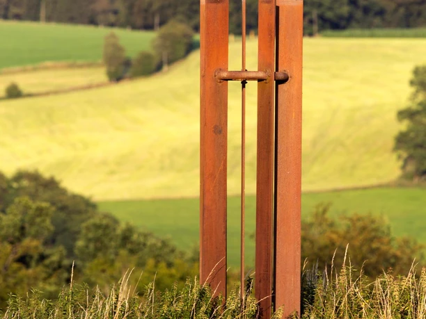 Friedens-Stele aus rostigem Stahl mit kleinem Vogel vor Wiesen und Wald
