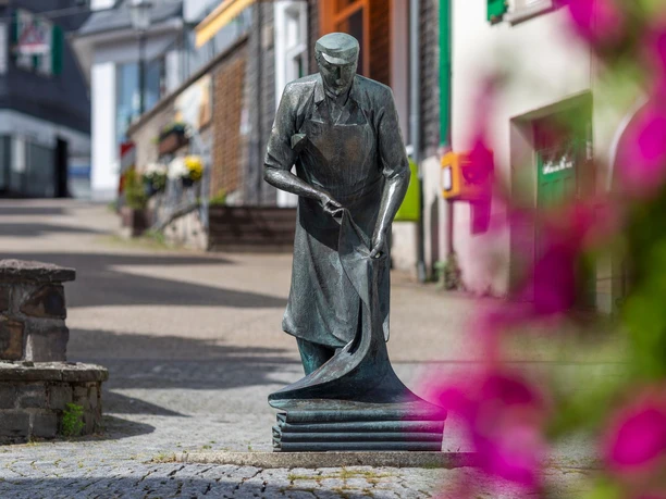 Bronzeskulptur Der Weber in Hückeswagen, rechts unscharfe pinke Blüten