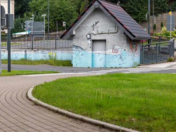 Bemaltes Regenüberlaufbecken mit Wasserhahn-Motiv, Geländer und Straße im Hintergrund