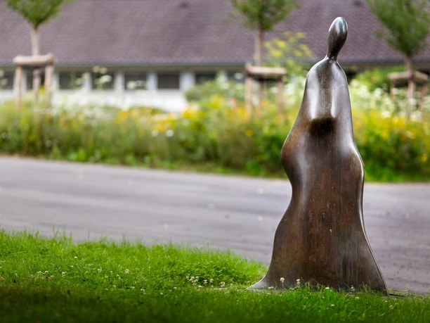 Bronzene Skulptur „Auf dem Weg“ auf einer Wiese vor Straße und Blumenbeet.