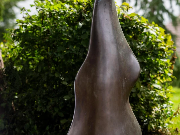 Bronzeskulptur „Auf dem Weg“ von Bernhard Guski vor grünem Strauchwerk