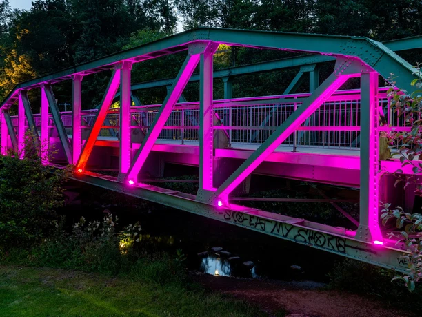 Pink beleuchtete Bogenbrücke über der Wupper, umgeben von dichtem Grün